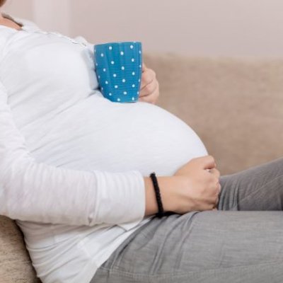A woman in a white top reclines on a couch, resting a blue coffee cup on her pregnant stomach